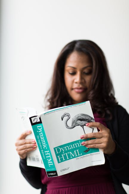 A woman with dark hair, dressed in a dark blazer and deep pink top, is holding and reading a book titled 'Dynamic HTML' by O'Reilly, featuring an illustration of a flamingo on the cover. She stands ag