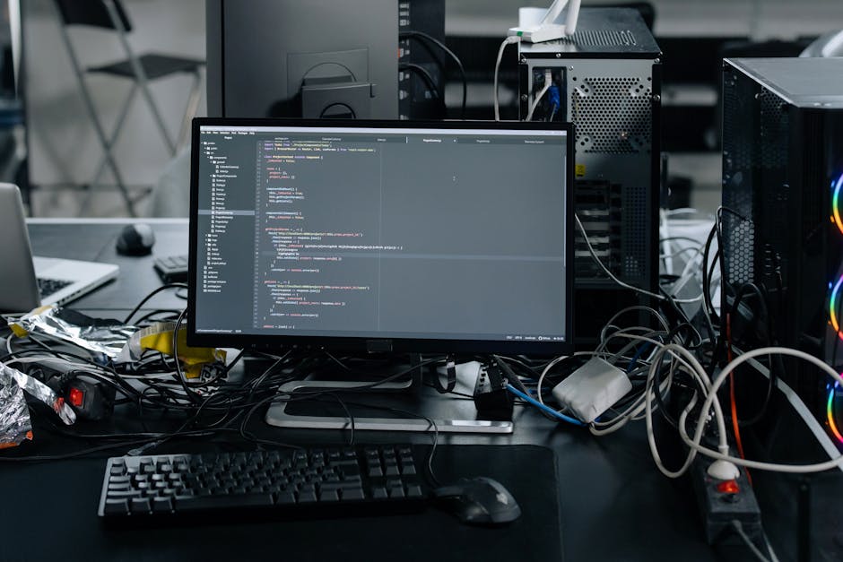 A cluttered workspace featuring a black desk with a computer monitor displaying lines of code, accompanied by a keyboard and mouse. Surrounding the monitor are various cables, some loosely tangled, co